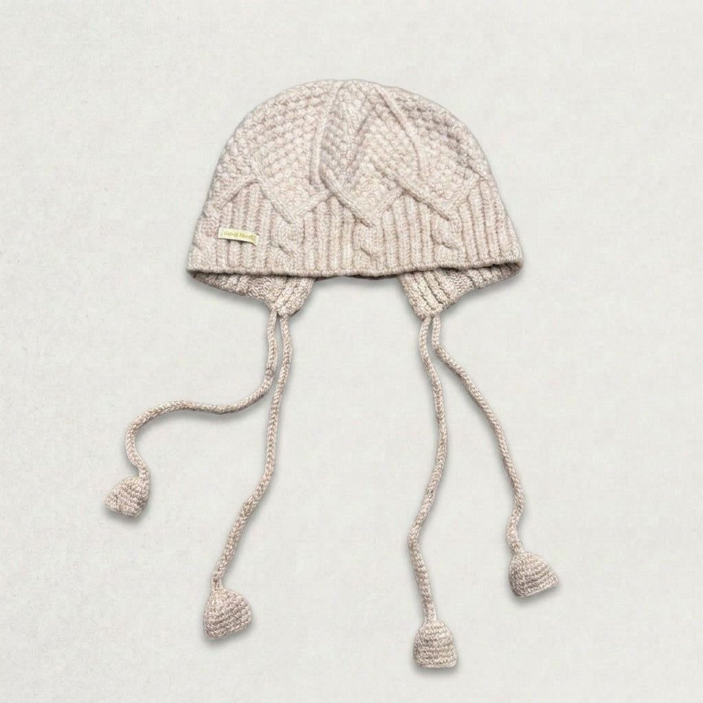 Beige cable-knit jellyfish hat with four dangling cords, laid flat