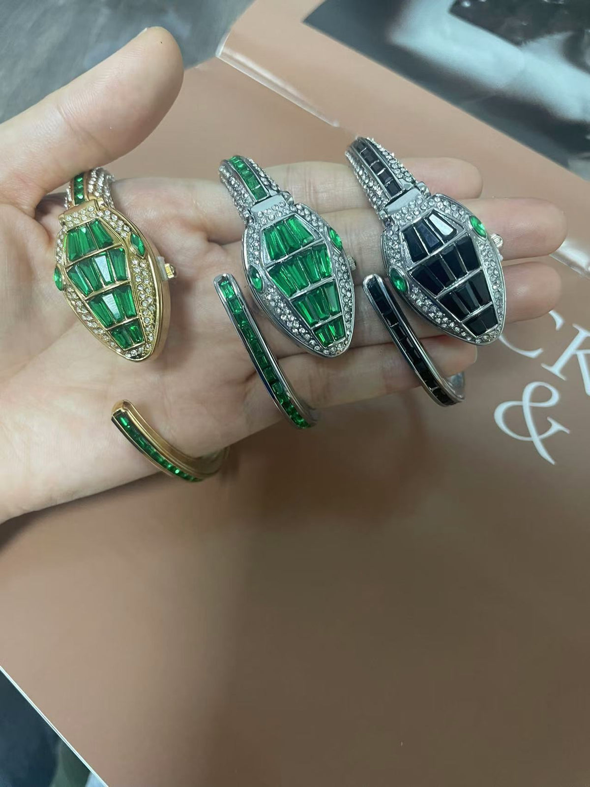 Three snake bangle watches in gold green, silver green and silver black held in a palm