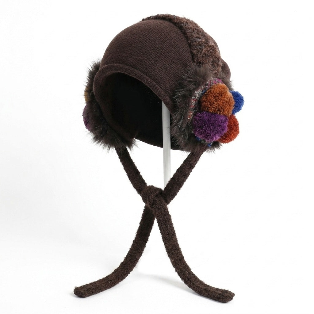 Brown ear-flap hat with matching ties and purple, orange and blue pom-poms, on a stand