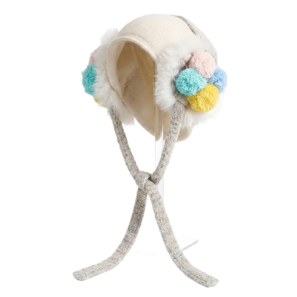 White ear-flap hat on a stand, showing pastel pom-poms and grey knitted ties
