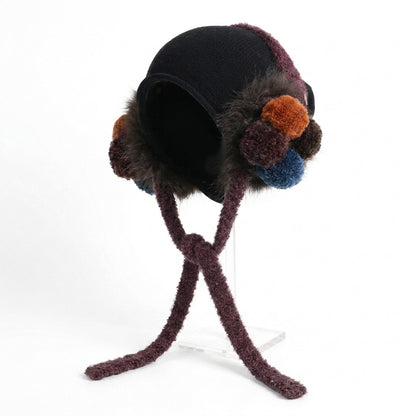 Black ear-flap hat with plum trim and ties, with orange and blue pom-poms, on a stand