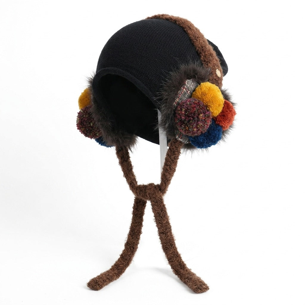 Black ear-flap hat with brown trim and ties, with yellow, red and blue pom-poms, on a stand