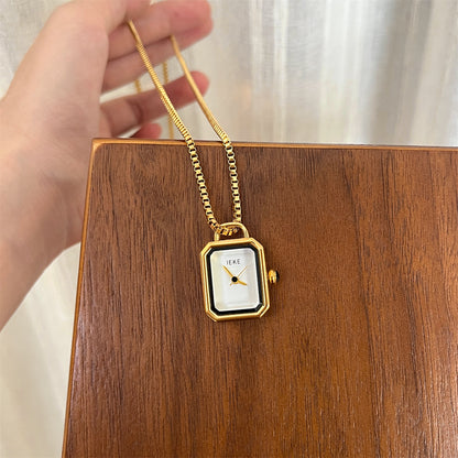 Gold rectangle pendant watch with a white dial held over a wooden board