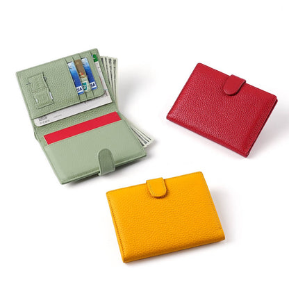 Three textured leather bifold wallets in sage green, red, and mustard yellow, arranged on a clean white background. The green wallet is open, showing card slots, a cash compartment, and a red interior stripe.